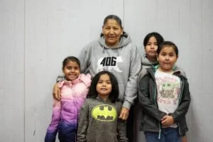A group of five people, likely a family, stand together in front of a plain wall.