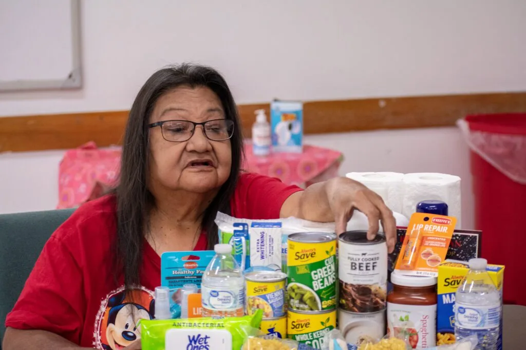 A person is reaching for canned food and supplies on a table. Items include canned beef, vegetables, and toiletries.