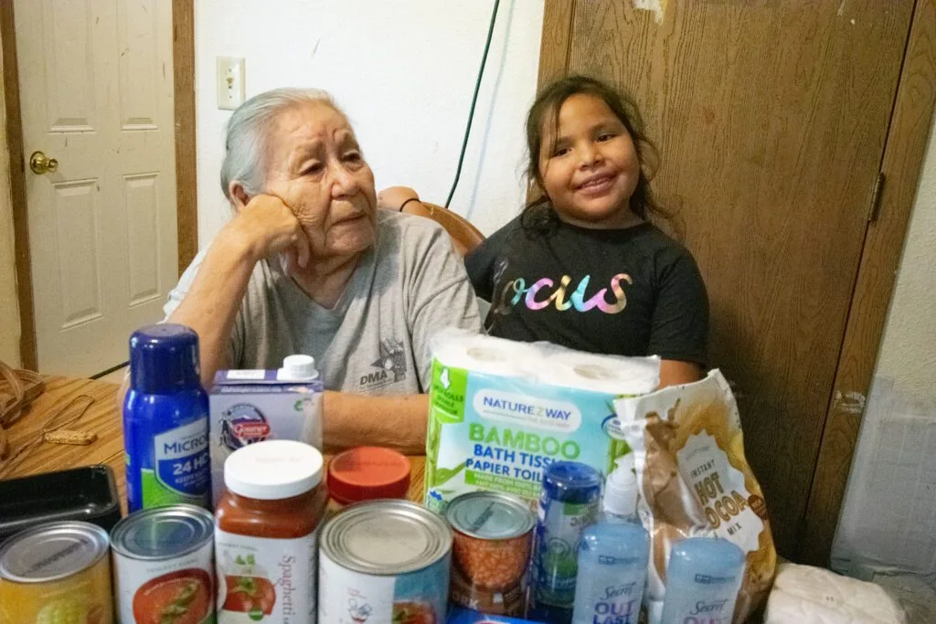 An elderly woman and a young girl sit at a table with various groceries and household items.