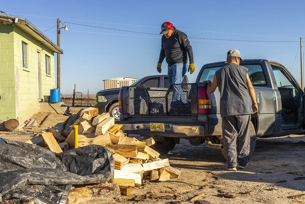 Two people unload firewood from a truck next to a house.