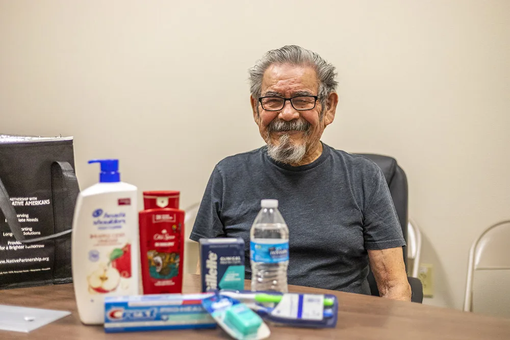 A smiling man sits behind a table with personal care items, including lotion and toothpaste. Southwest Indian Relief Council is relevant.