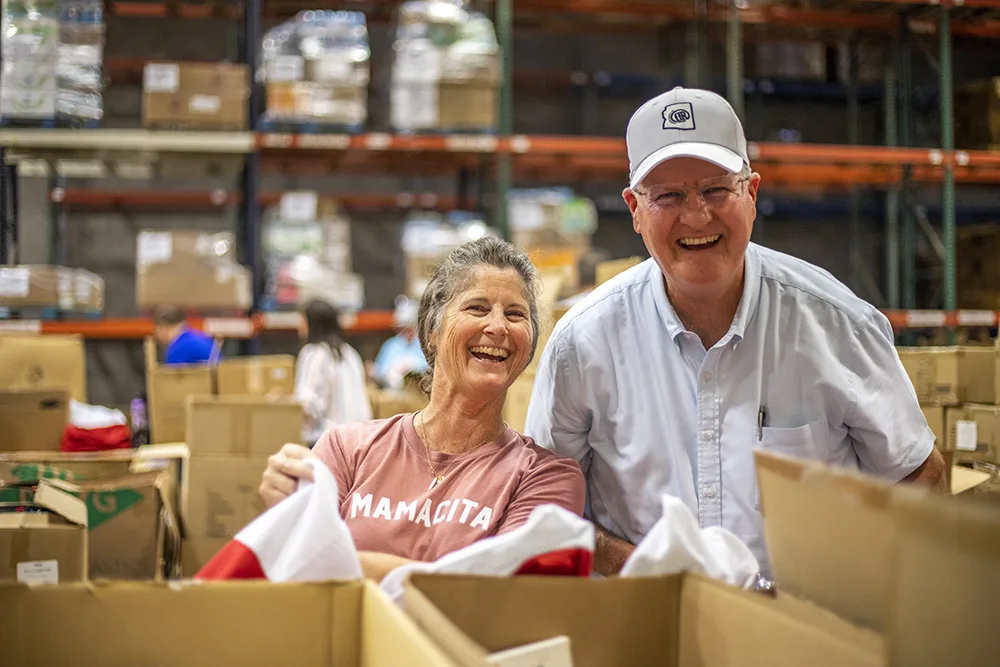 Two people are smiling while working in a warehouse or storage facility with boxes around them.