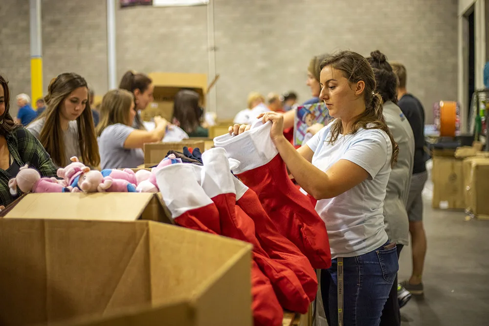 People are filling red stockings with toys in a warehouse setting.