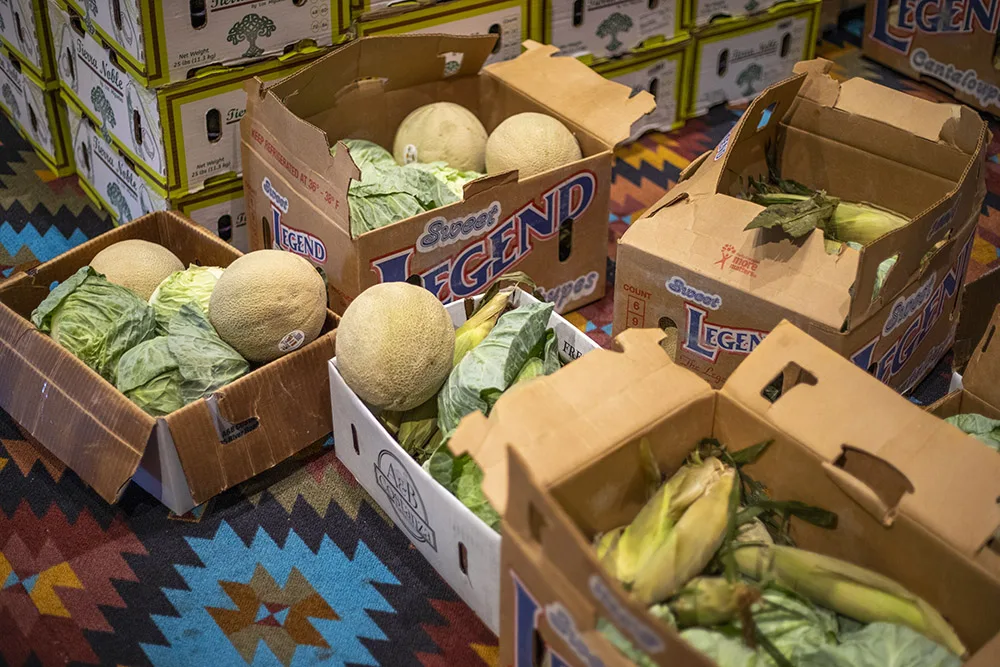 Boxes containing melons, cabbage, and corn are stacked on a colorful carpet.