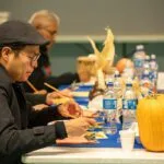 People are sitting at a table with food, drinks, and decorative pumpkins, engaged in conversation or dining.