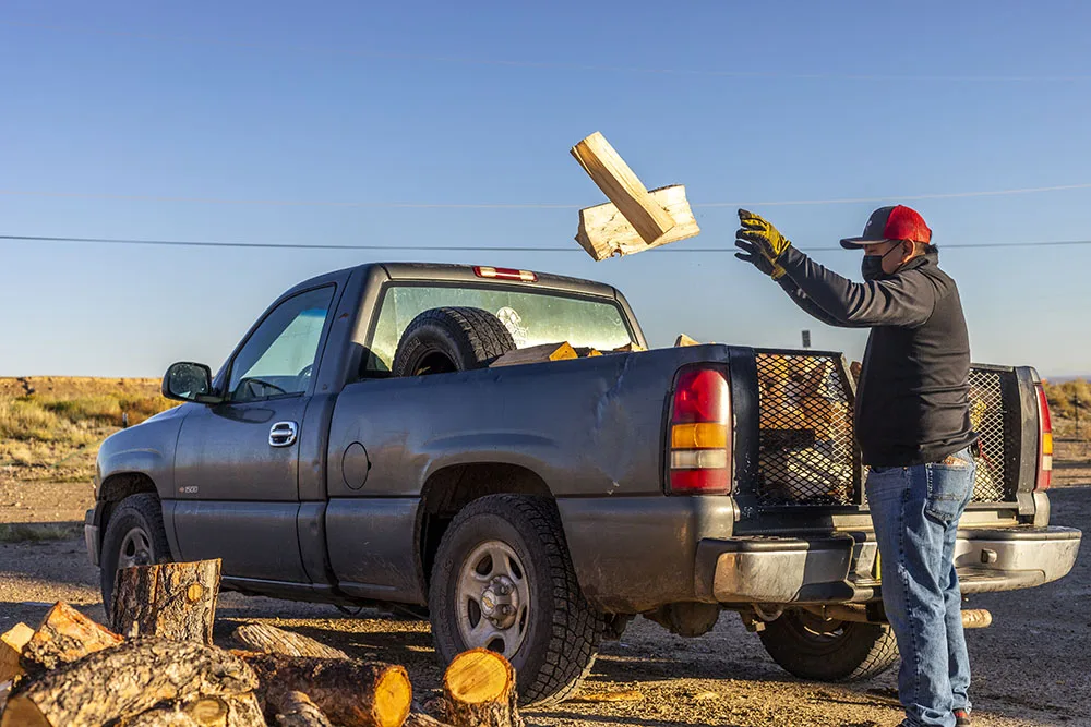 A person is tossing firewood into the back of a pickup truck in a rural setting.