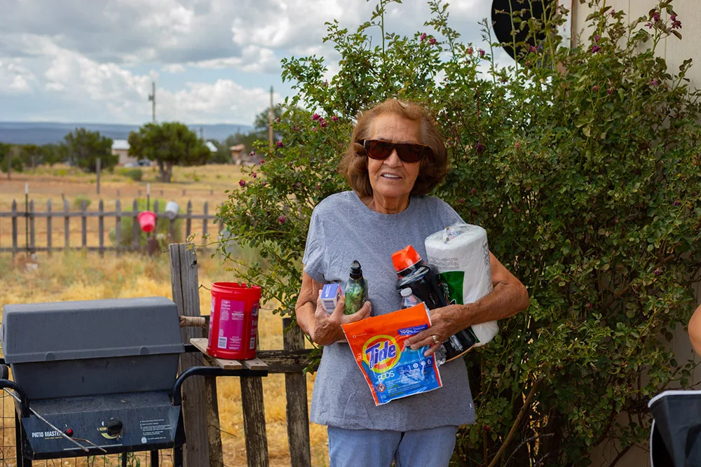 A smiling woman outdoors holding household supplies and a cleaning product package, possibly associated with the Southwest Indian Relief Council.