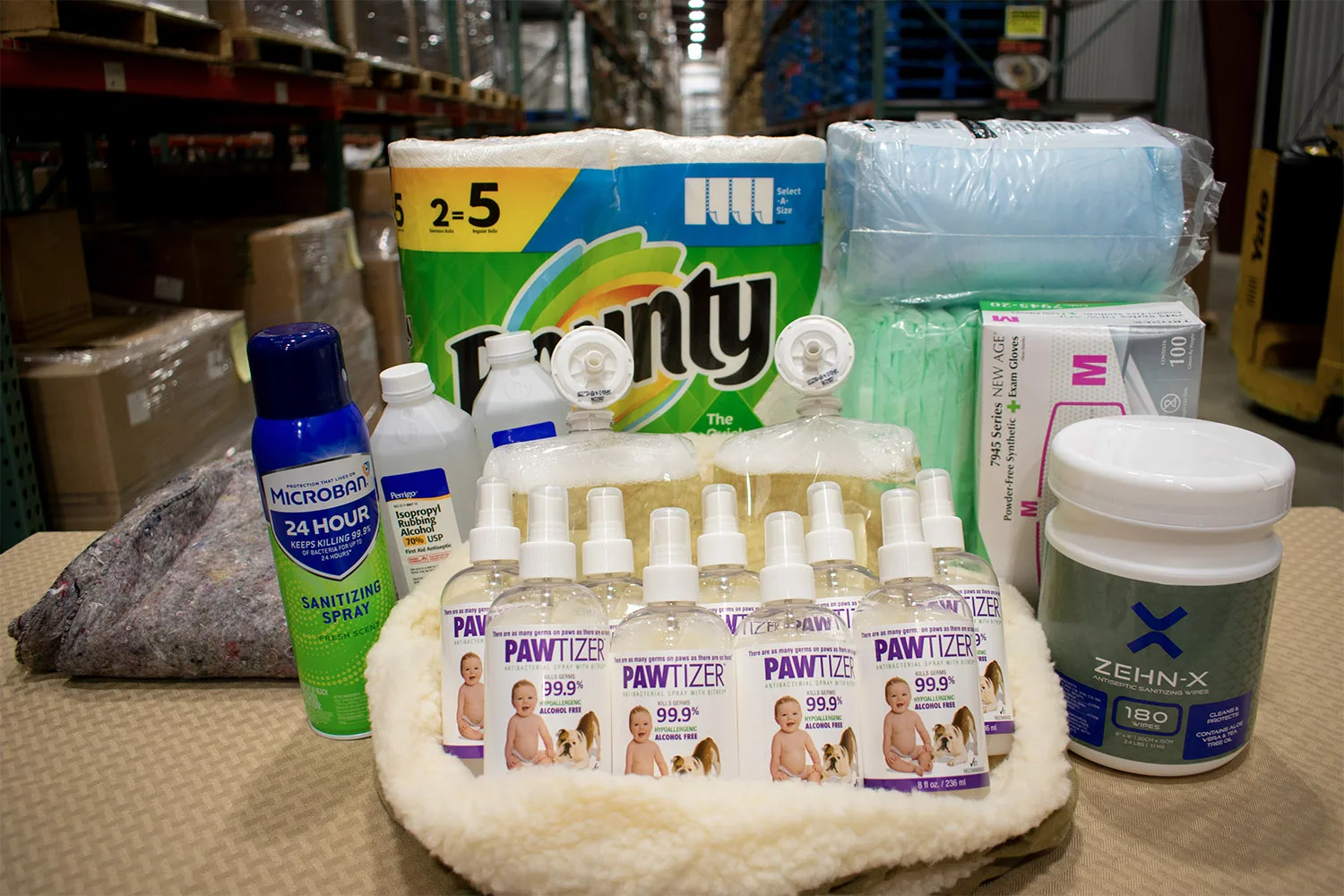 Sanitizing products, paper towels, gloves, and cleaning supplies are arranged on a table in a warehouse setting.