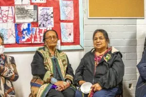 People are sitting together indoors near a bulletin board with colorful artwork.