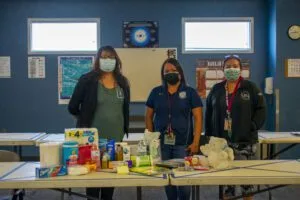 Three people wearing masks stand behind a table with health and hygiene supplies.