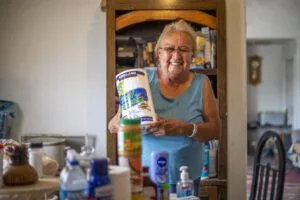 A person is smiling and holding household supplies, standing in a room with various items on a table.