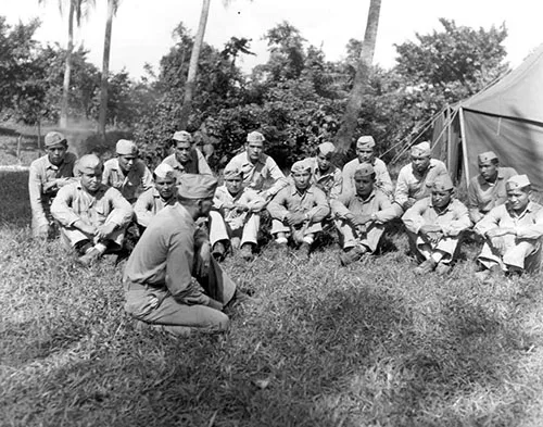 A group of soldiers in uniform is gathered on grass, possibly during a briefing or meeting outdoors.