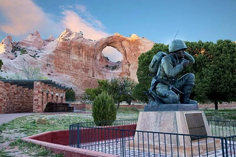 A statue of a soldier kneeling, located in a desert landscape with a natural arch in the background. Navajo Code Talkers memorial.